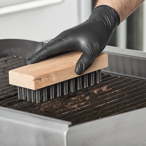 A person wearing black gloves uses a Choice stiff steel bristle grill brush to clean a grill.