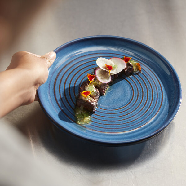 A hand holding a Libbey Canyonlands blue terracotta plate with a slice of meat and radish.