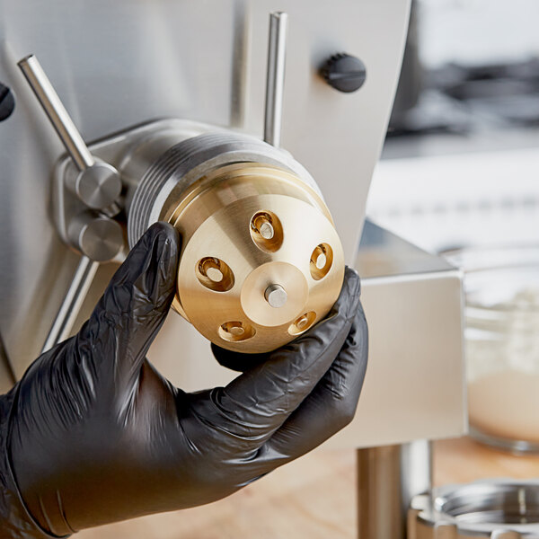 A person in black gloves using a gold Estella penne pasta die to grind a metal object on a counter.