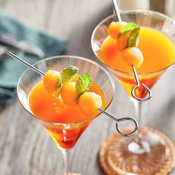 Two glasses of orange liquid with fruit on stainless steel skewers.