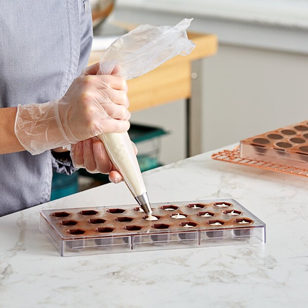 A woman using a pastry bag to fill a Pavoni hexagon candy mold with chocolate.