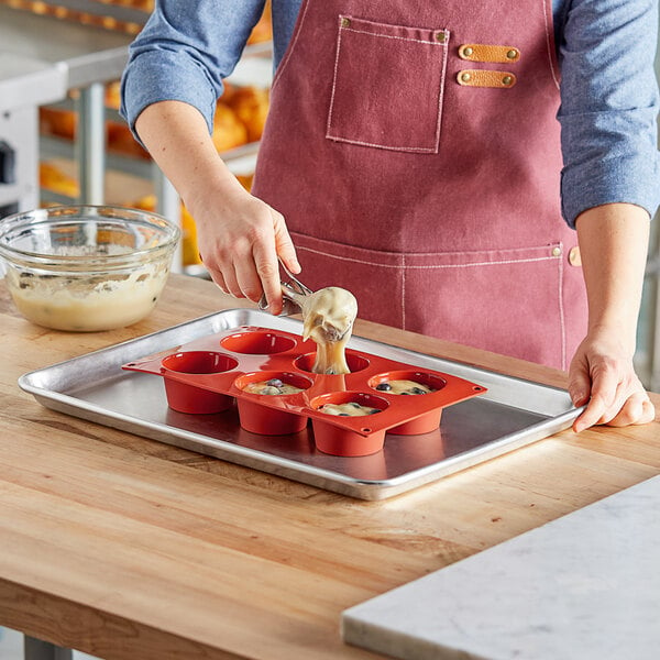 A woman in an apron using a Pavoni Formaflex silicone muffin tin to pour liquid into a bowl of white batter.