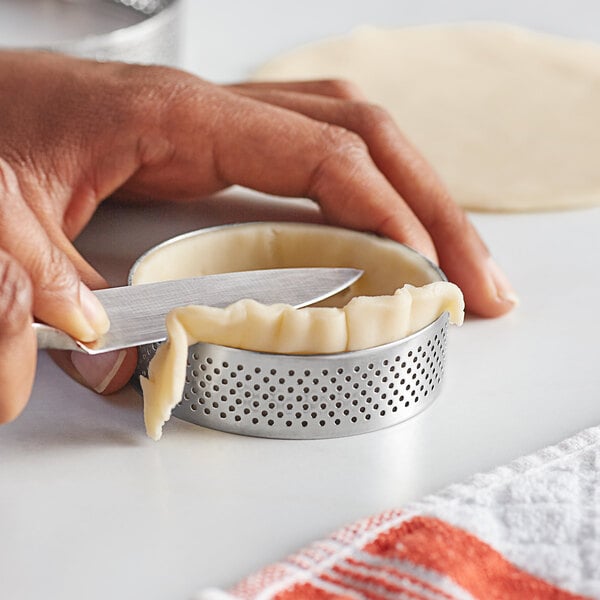 A person using a knife to cut pastry dough in a stainless steel tart ring.