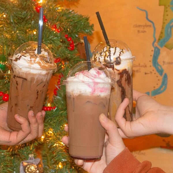 A group of three plastic cups filled with iced chocolate drinks, each topped with whipped cream and a black straw, held up in front of a decorated Christmas tree.