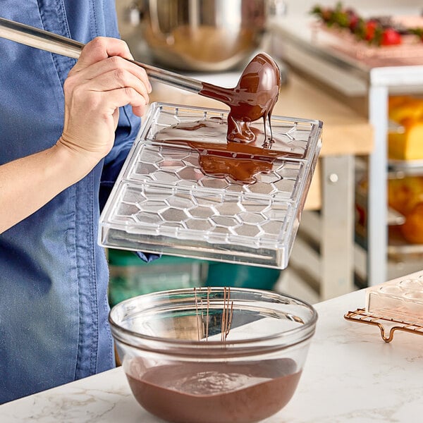 A hand pouring chocolate into a Pavoni Polycarbonate chocolate mold.