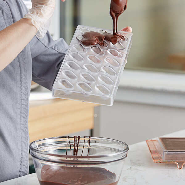 A person pouring chocolate into a Pavoni Praline oblong candy mold on a counter.