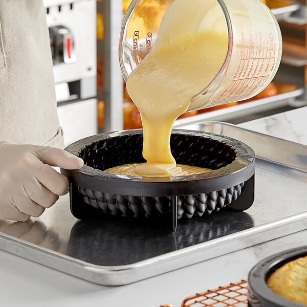 A person pouring yellow liquid from a glass measuring cup into a black Pavoni Pavocake Facon silicone baking mold.