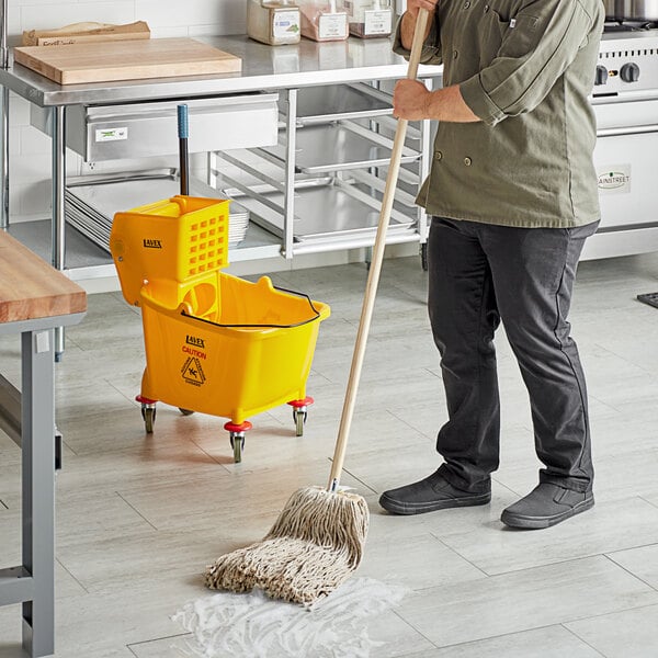 A man in a green jacket mopping a white floor with a Lavex wet mop and yellow bucket.