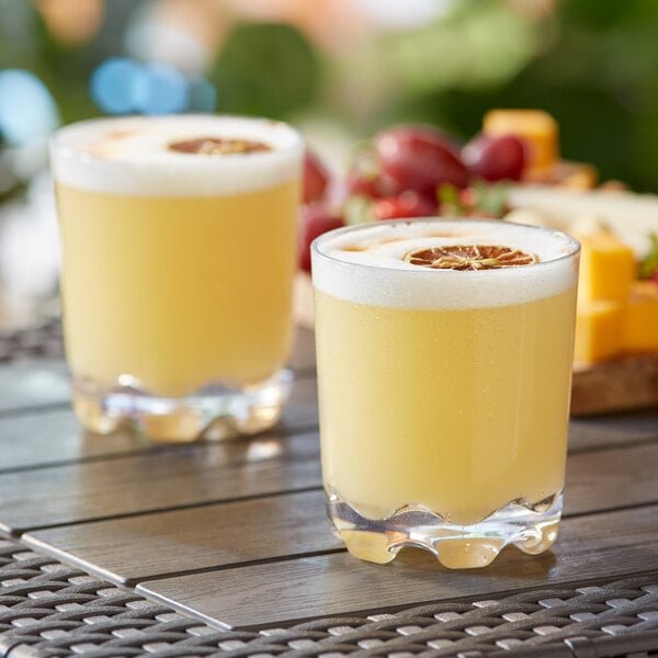 Two Acopa Endure Tritan plastic rocks glasses filled with yellow liquid and orange slices on a table.