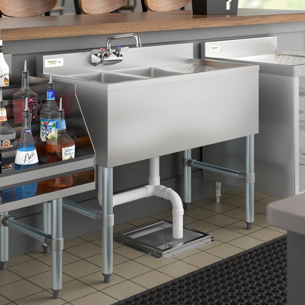 A Regency stainless steel underbar sink with right drainboard and faucet on a counter with bottles of liquor.