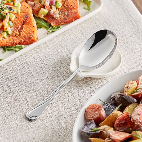 An Acopa stainless steel serving spoon on a plate of food next to a fork and another plate of food.