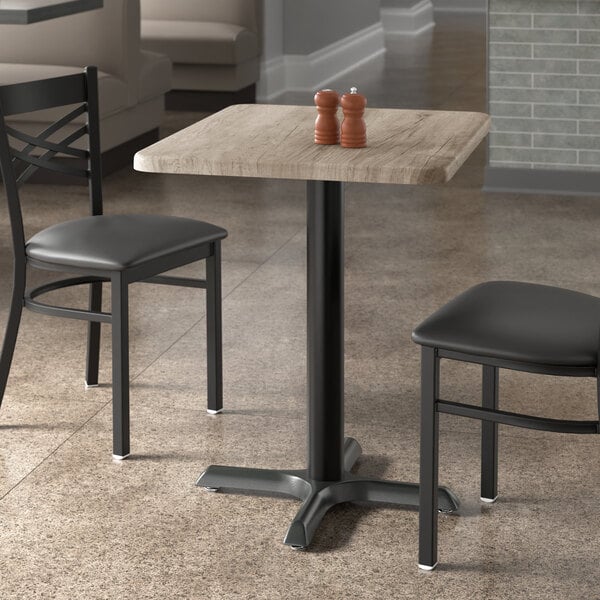 A rectangular standard height table with a gray wood finish and a black metal base, accompanied by two black chairs.