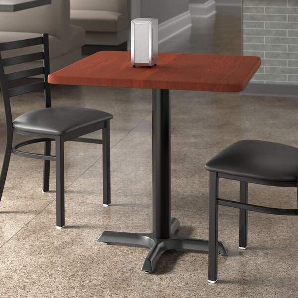 A Lancaster Table & Seating red mahogany thermo-formed MDF table top with a rectangular object on it and two black chairs.