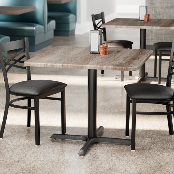 A square standard height table with a gray wood finish and black metal base, set in a restaurant setting.