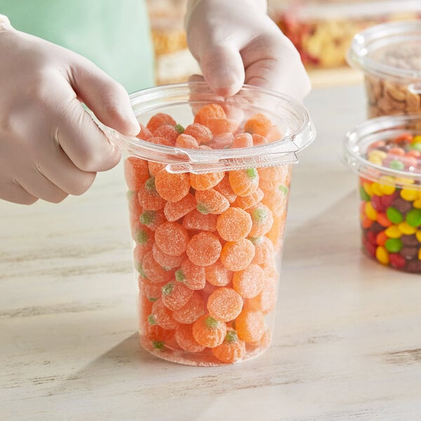 A gloved hand holding a Inline Plastics Safe-T-Fresh plastic container filled with orange candies.