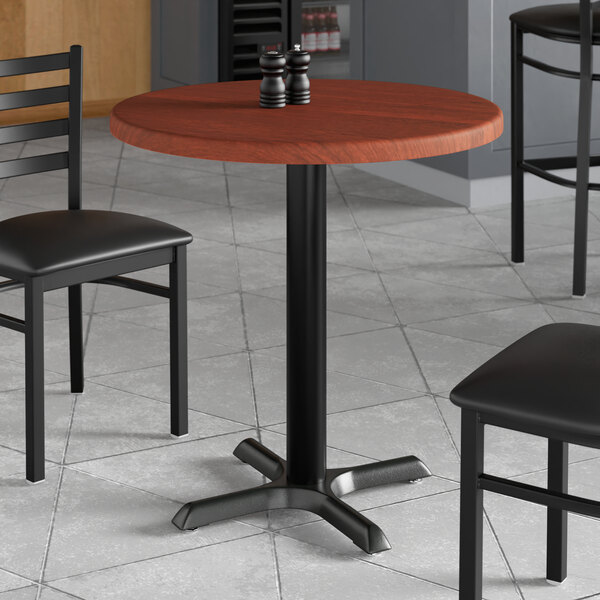 A Lancaster Table & Seating round table with a red mahogany finish and black chairs around it.