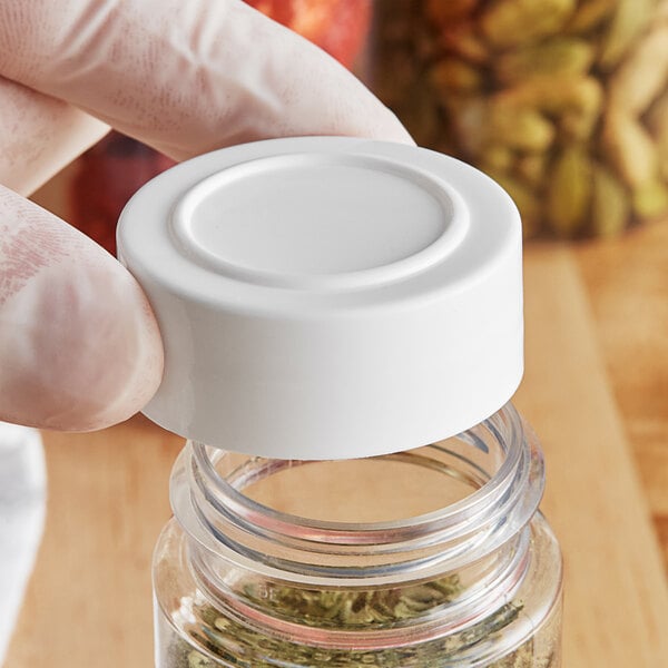 A hand holding a white 43/485 unlined polypropylene cap over a small jar of herbs.