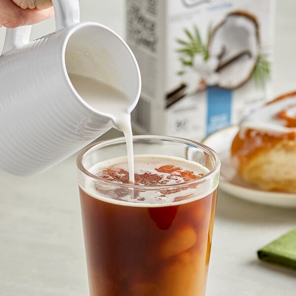 A person pouring So Delicious Organic Vanilla Coconut Milk into a glass of brown liquid.