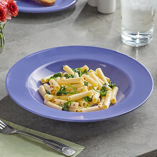 A plate of pasta in an Acopa purple melamine bowl with a silver spoon and a white cup on a green table.