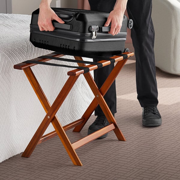 A person holding a black suitcase on a Lancaster Table & Seating walnut wood folding luggage rack.