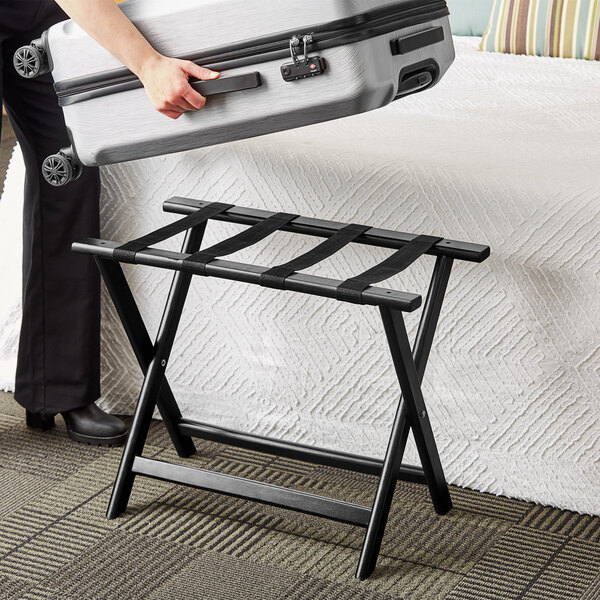 A woman uses a Lancaster Table & Seating black wood folding luggage rack to hold a suitcase.