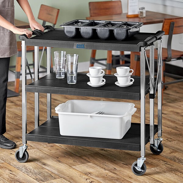 A woman standing next to a black Lavex plastic utility cart with a tray of dishes.