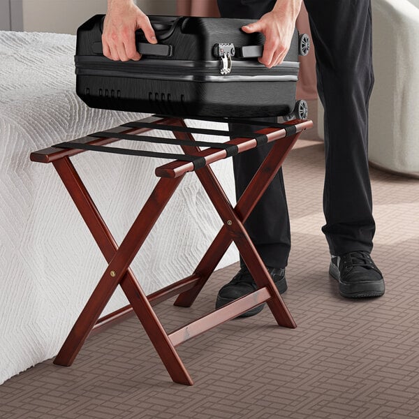 A person holding a suitcase on a Lancaster Table & Seating mahogany wood folding luggage rack.