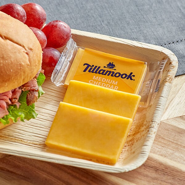 A plate with a sandwich, apple, and grapes with a Tillamook Medium Yellow Cheddar cheese snack portion.