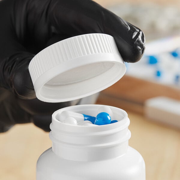 A hand in a black glove opening a white plastic bottle of blue and white pills with a white child resistant cap.