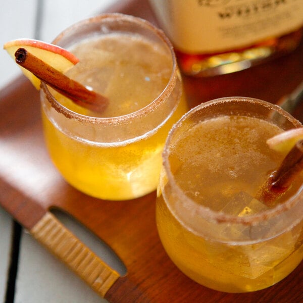 Two Tossware plastic Vino Jr. glasses of apple cider with cinnamon sticks and an apple on a wooden tray.