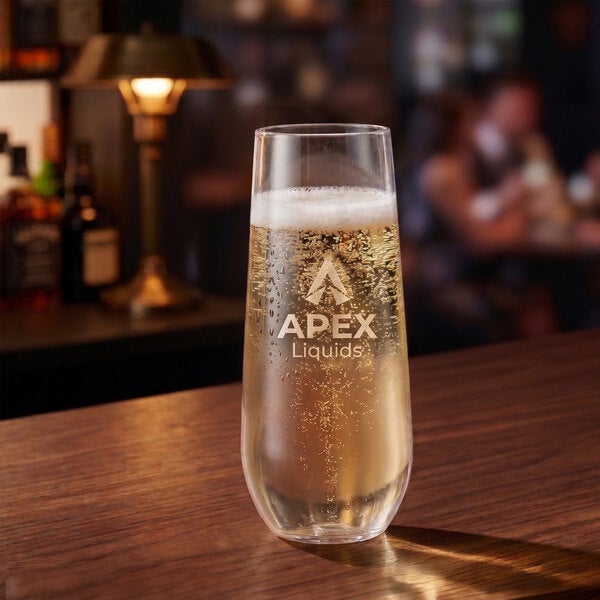A clear plastic champagne flute glass with customizable branding, filled with a sparkling beverage, placed on a wooden bar counter.