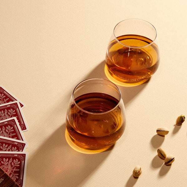 Two Tossware rocks glasses filled with brown liquid next to a deck of cards and pistachios.
