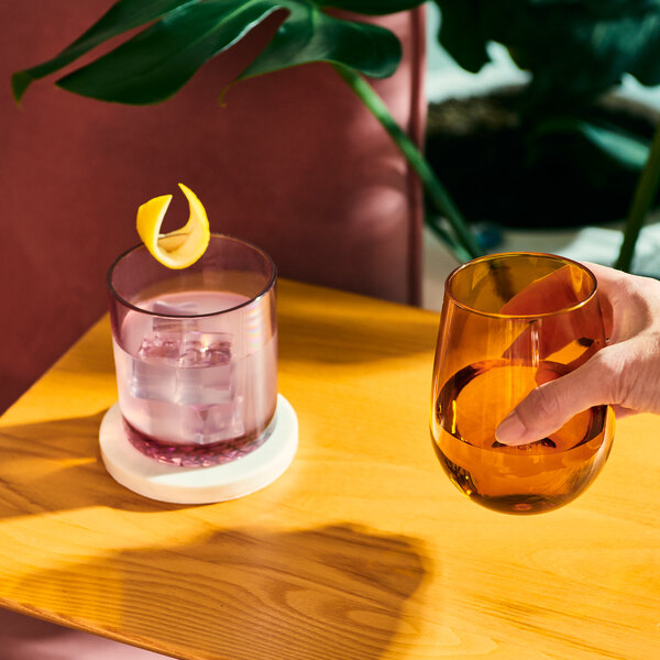 A hand holding a Tossware Reserve Go-To Blush Tritan plastic rocks glass of liquid with a lemon slice.