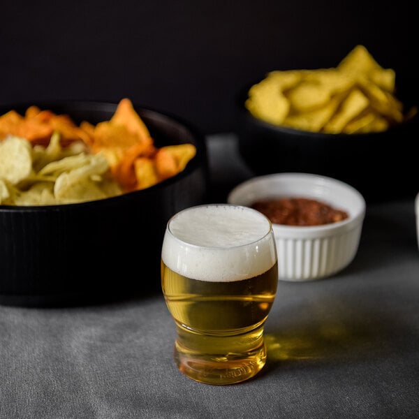 A Tossware plastic taster glass of beer next to a bowl of chips.