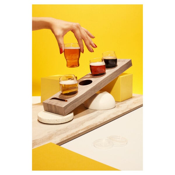 A hand holding a Tossware POP Taster glass of beer on a wooden flight tray with 3 empty spots.