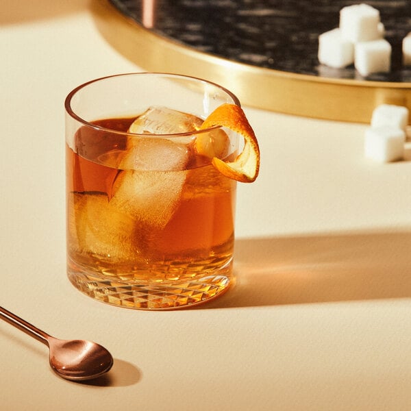 A Tossware tritan plastic rocks glass of whiskey with ice and an orange slice.