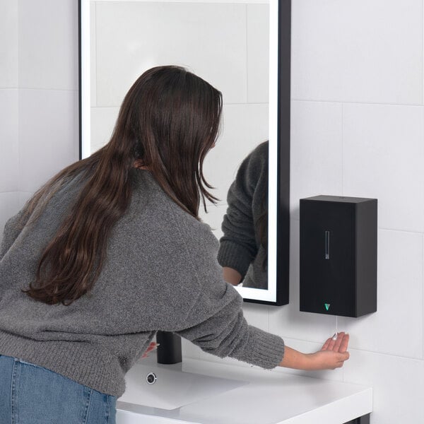 A person is using a wall-mounted, black automatic soap dispenser next to a bathroom sink and mirror.