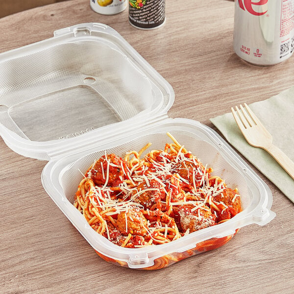 A Genpak clear plastic hinged container with spaghetti and meatballs inside on a table with a wooden fork and a cup of soda.