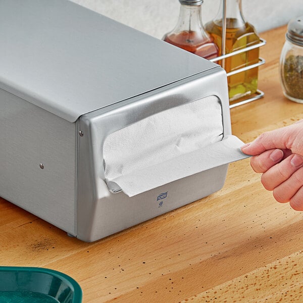 A hand pulling a white Tork dispenser napkin out of a silver box.