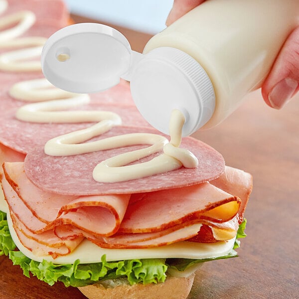 A person pouring white mayo from a bottle onto a sandwich.