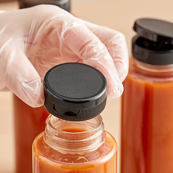 A hand wearing a glove is placing a black plastic flip top lid onto a bottle filled with orange liquid.