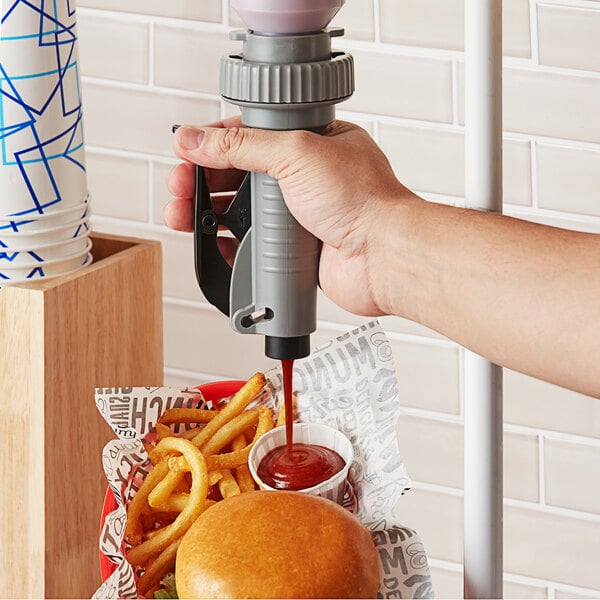 A hand holding a grey Asept condiment pump with red liquid over a table with a burger and fries.