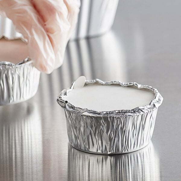 A hand in a plastic glove putting a white lid on a round aluminum Baker's Lane foil container.