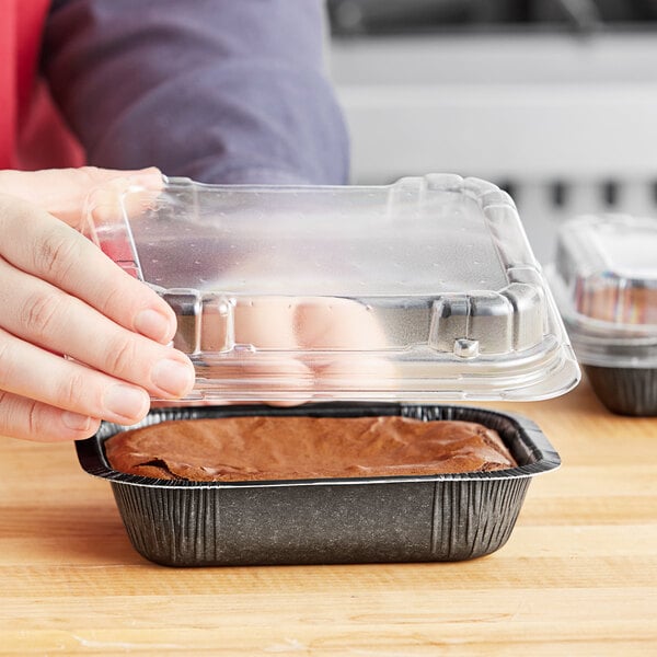 A person holding a Solut clear plastic lid over a brownie in a black container.