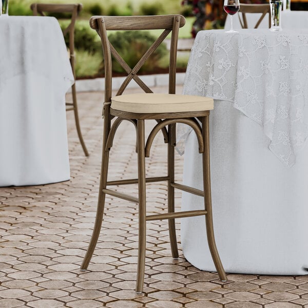 A Lancaster Table & Seating outdoor cross back bar stool with a tan cushion on the seat.