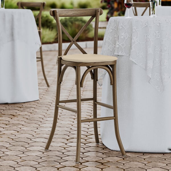 A Lancaster Table & Seating Vineyard Series Whiskey Brown outdoor cross back bar stool with a Chardonnay tan linen cushion on a patio table.