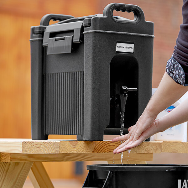 A person washing their hands in a black cooler filled with water using a CaterGator Sanitary Compliance label.