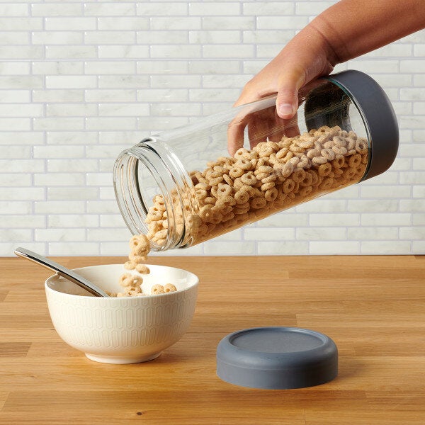 A clear glass jar with a gray threaded lid being used to pour cereal into a white bowl.