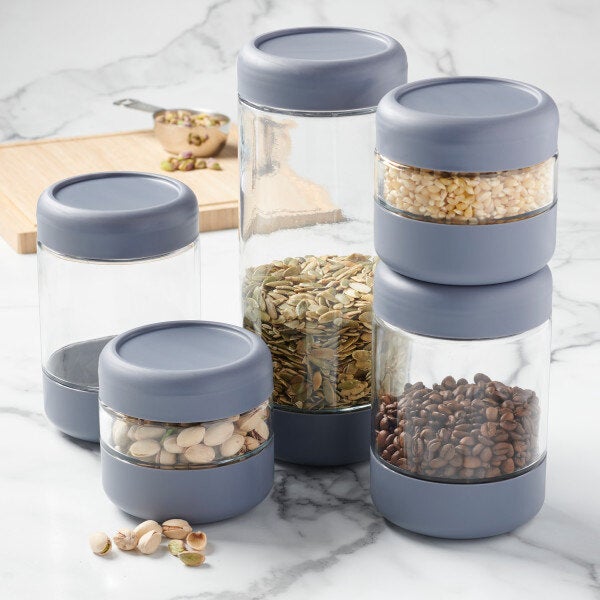 A set of stackable glass jars with gray threaded lids, each filled with various dry foods.