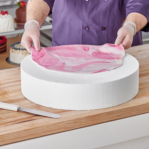 A person in a purple apron using a Baker's Lane foam round cake dummy to decorate a white and pink round cake.